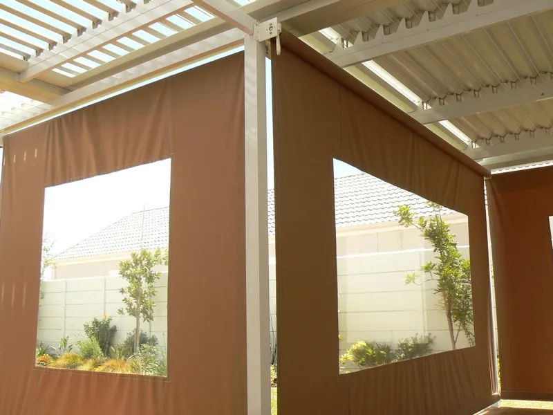 Taupe drop blinds with large clear PVC windows enclosing a patio under a white louvre and IBR combination roof, garden visible through the panels